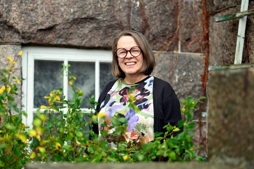 Anne taustallaan ikkuna ja kiviseinä, etualalla keltaisia kukkia
